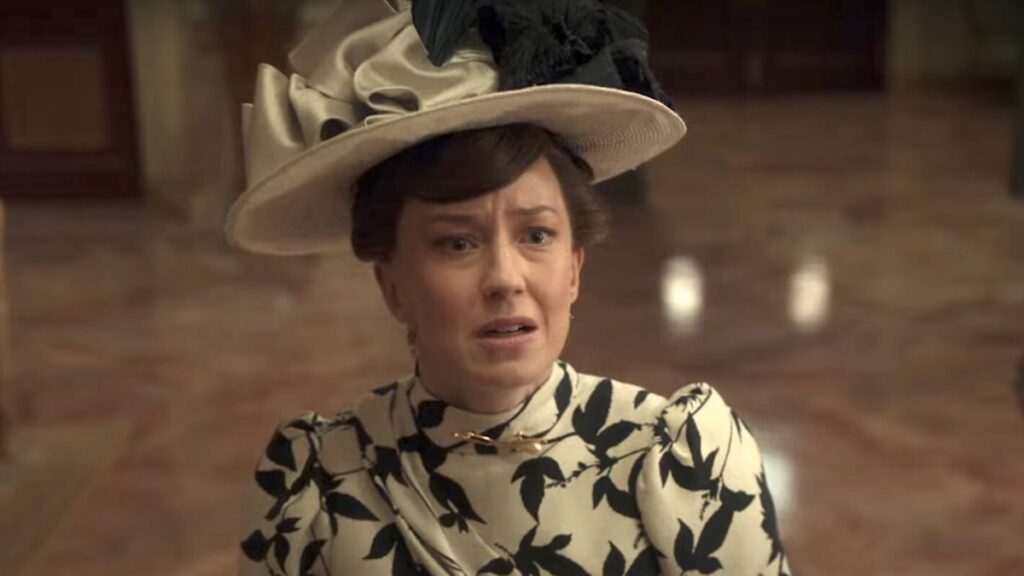 Carrie Coon as Bertha Russell in HBOs The Gilded Age, wearing a wide brimmed hat in a marble hallway